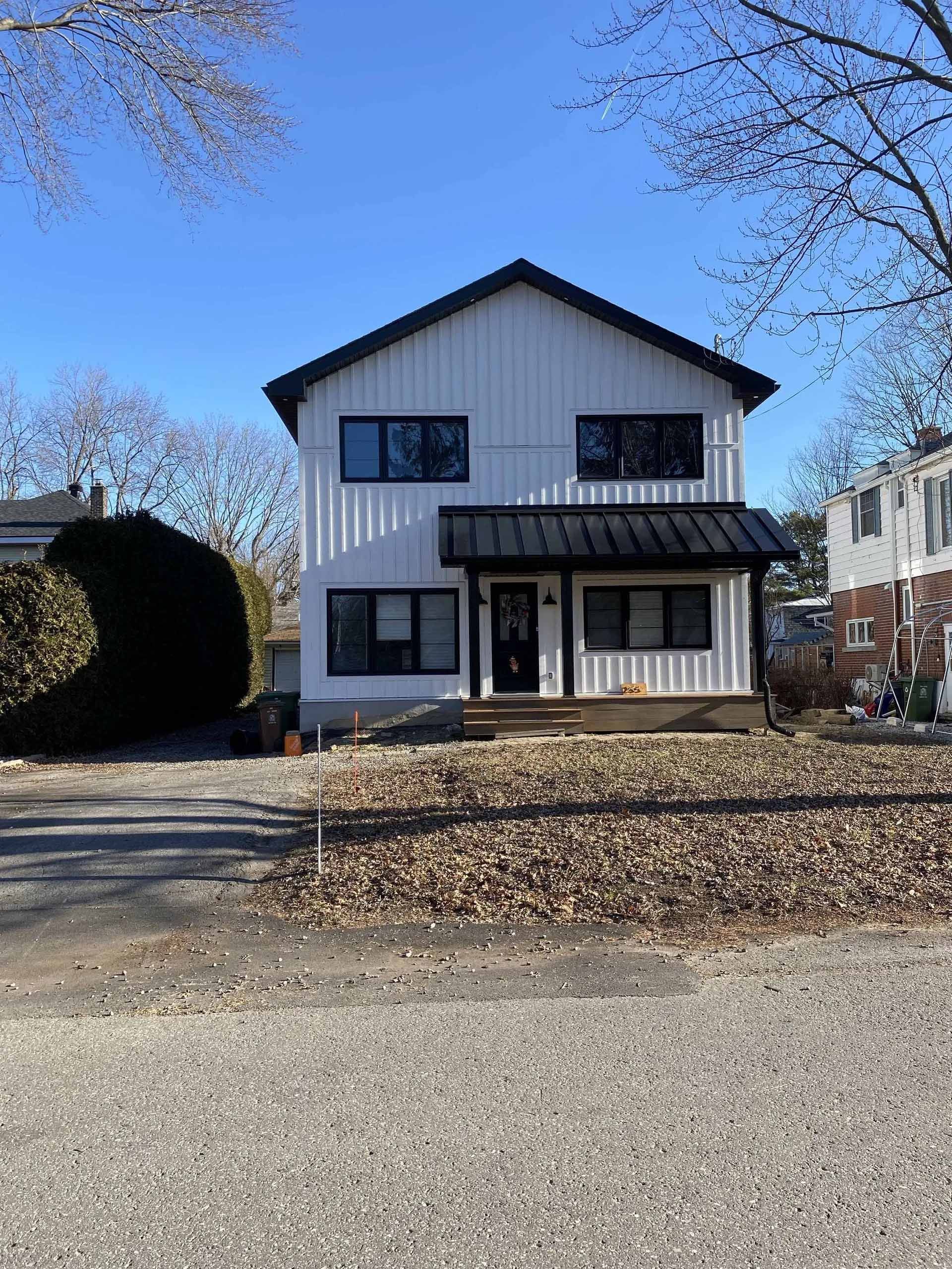 Maison blanche à deux étages avec toit et boiseries noires, porche, fenêtres. Allée de gravier, arbres dénudés, ciel bleu.