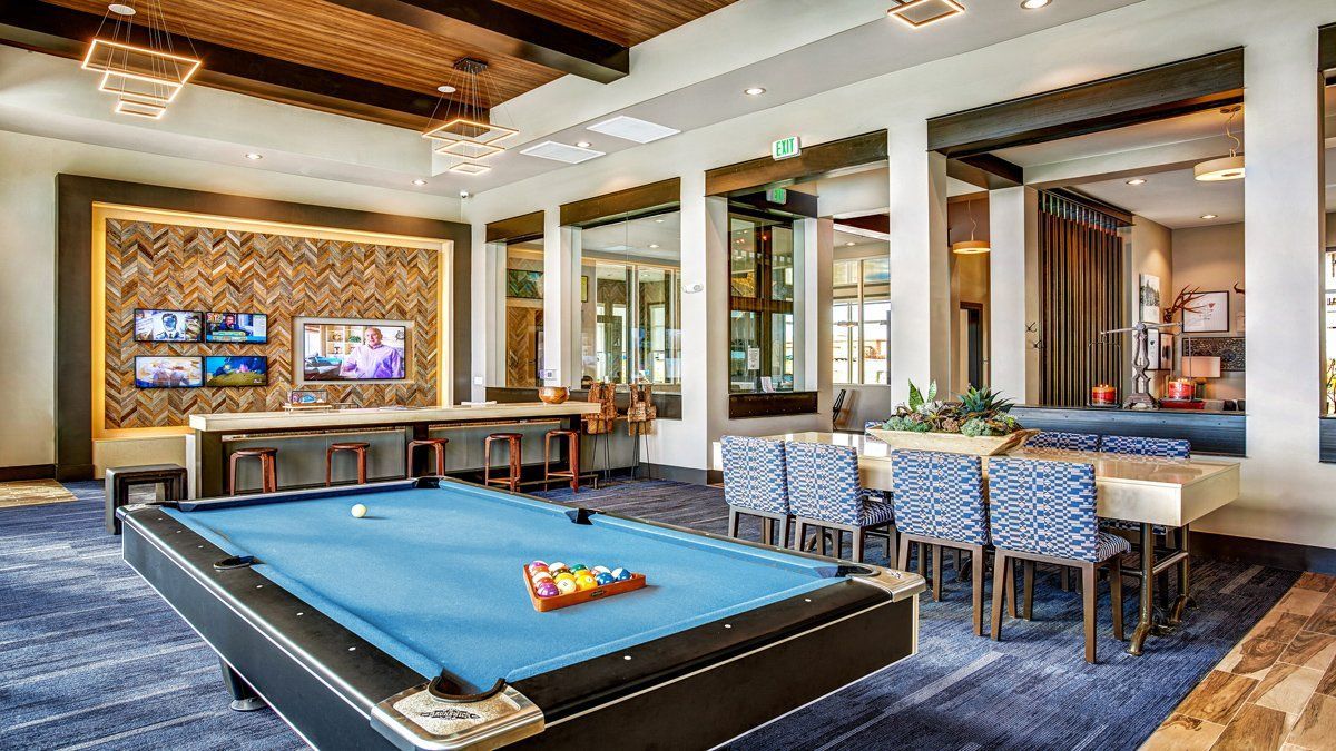Pool table in a lounge area, with a TV, table, and chairs. Bright space with blue carpet.