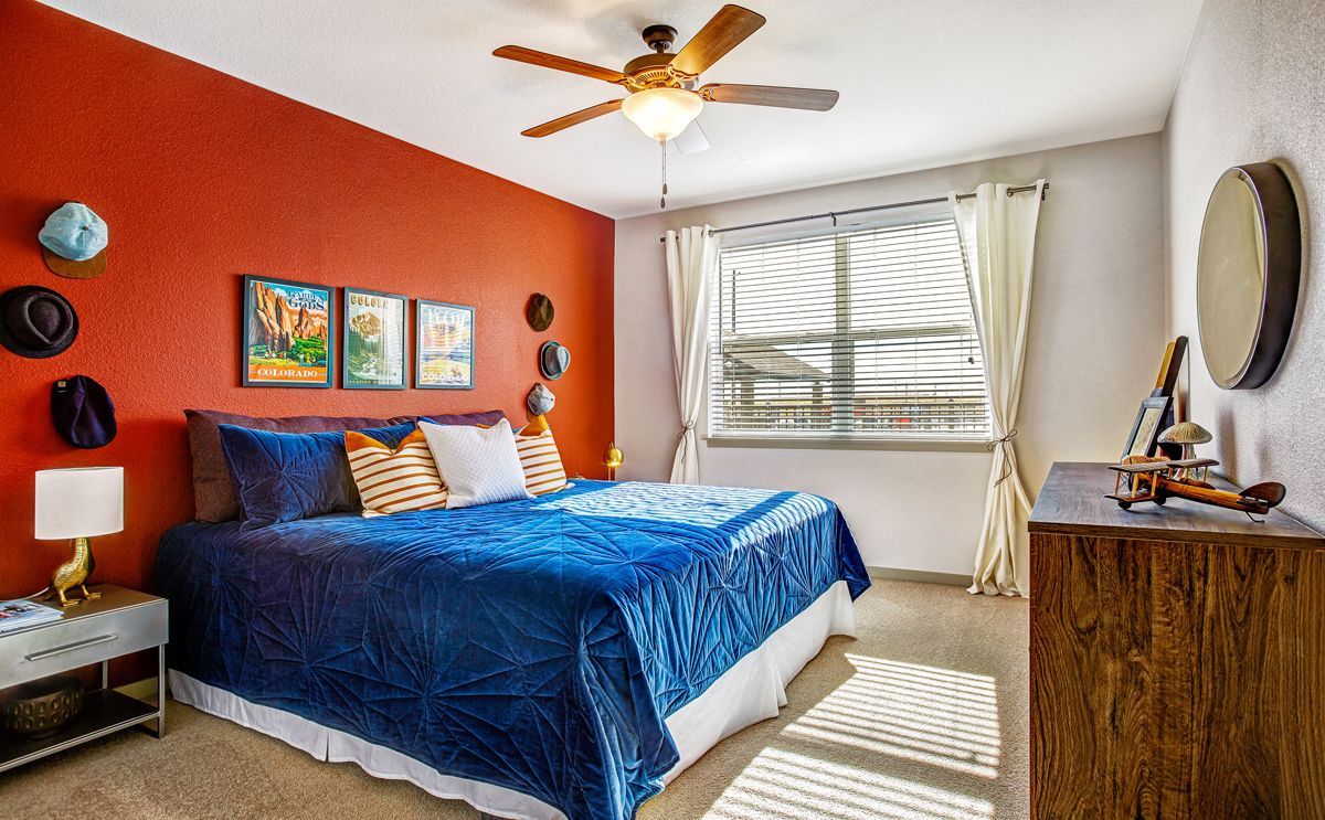 Bedroom with red accent wall, blue bedspread, hats, art, and wooden dresser near a window.