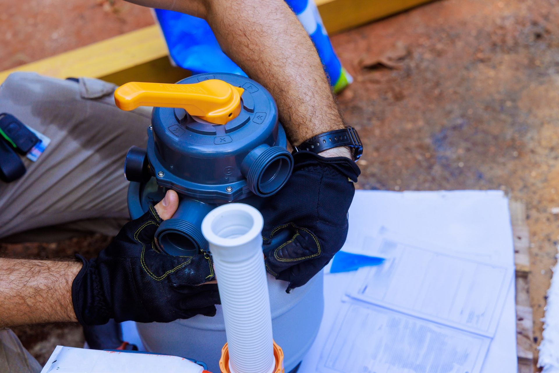 Person installing a pool filter, wearing gloves, holding a pipe, orange handle, outdoors.