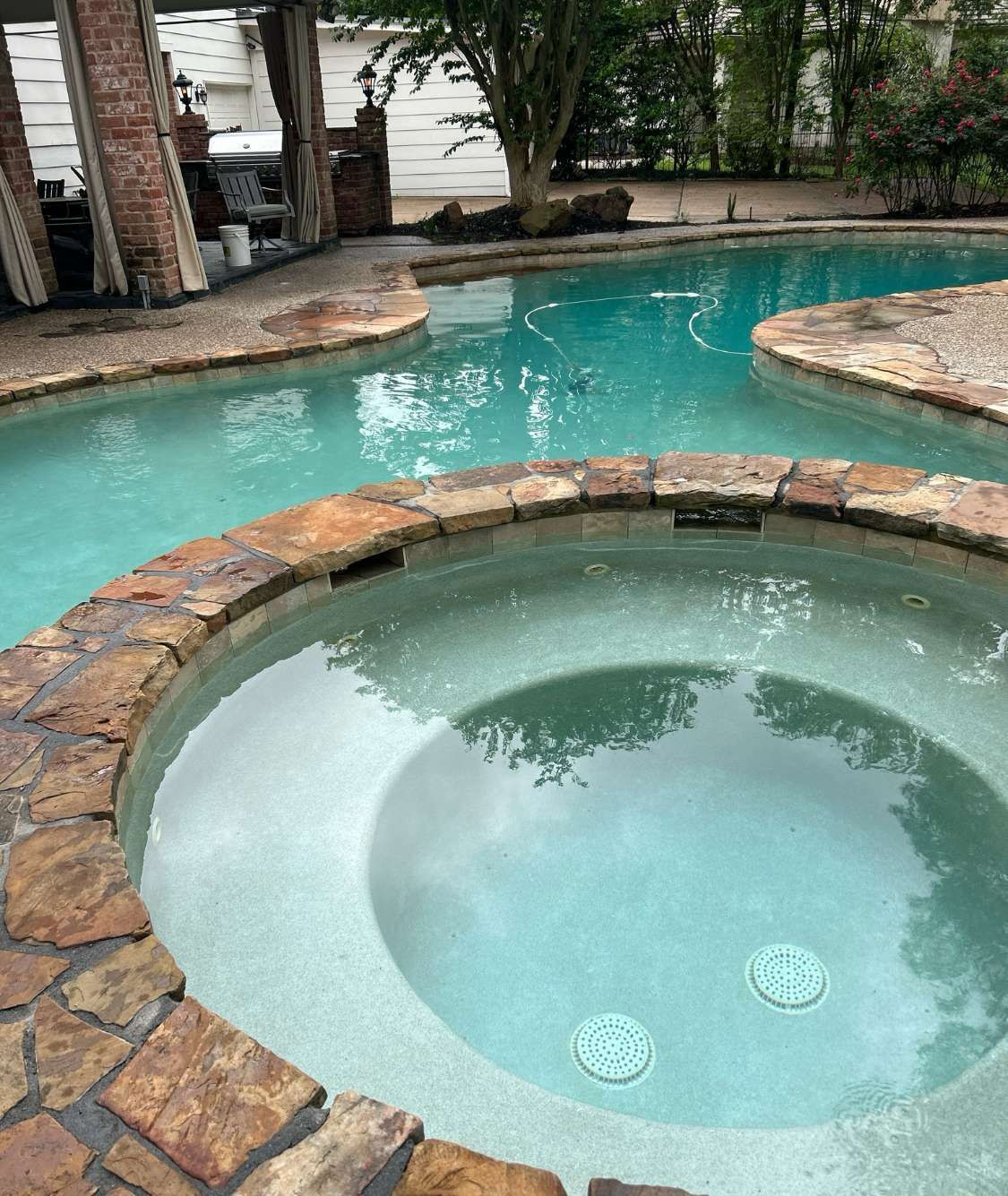 Stone-edged pool and hot tub filled with turquoise water. A shaded structure and trees are visible in the background.