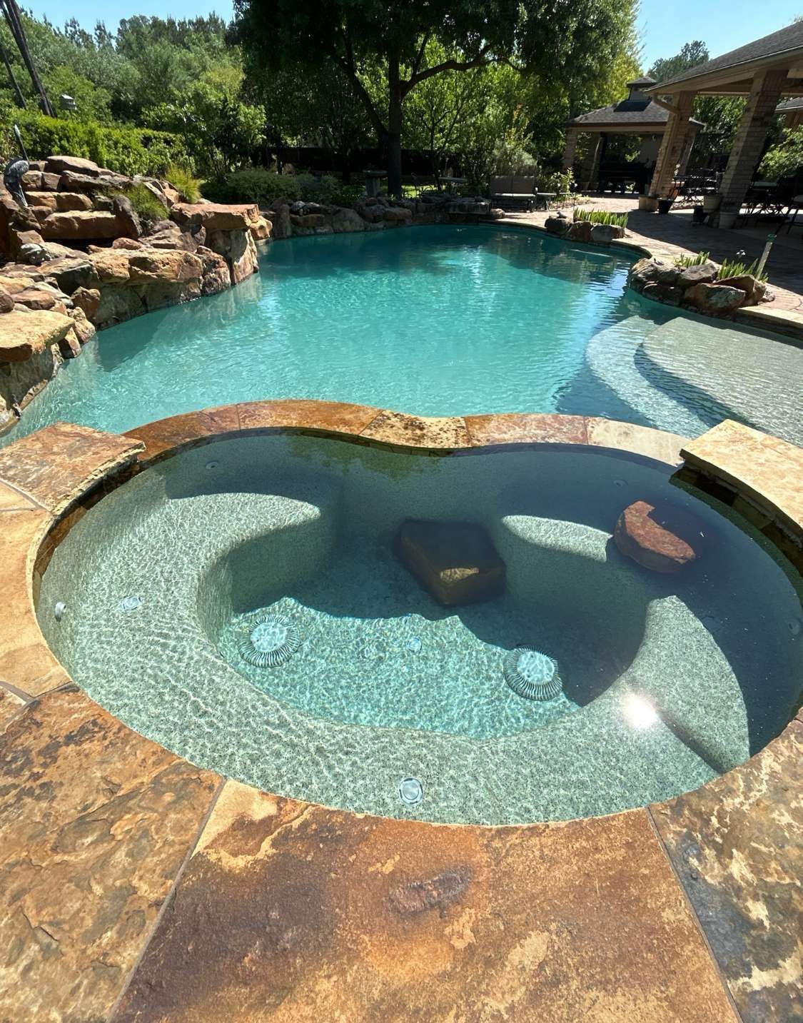 Swimming pool with attached spa, stone deck, and surrounding landscaping. Turquoise water.