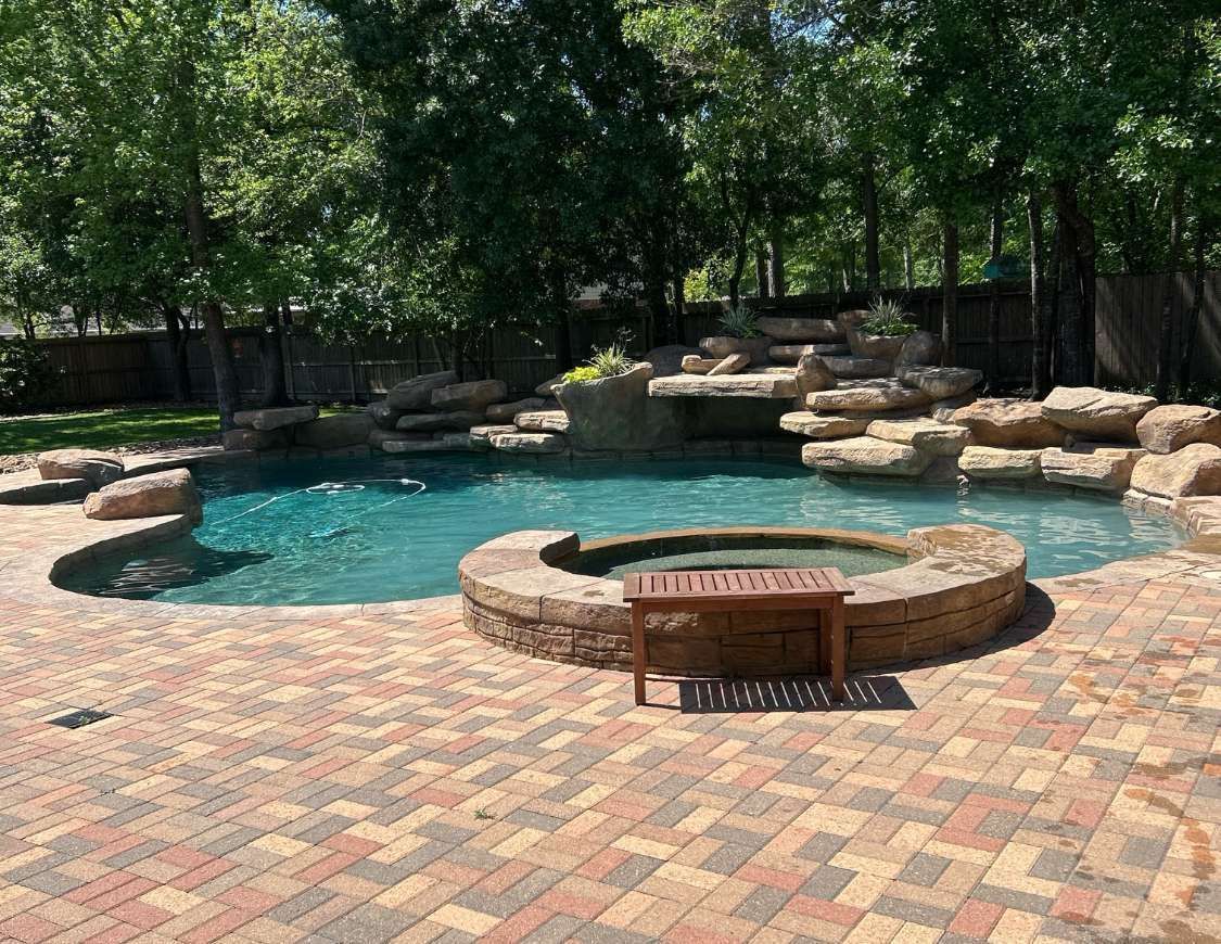 Backyard pool with rock waterfall, spa, and brick patio surrounded by trees.
