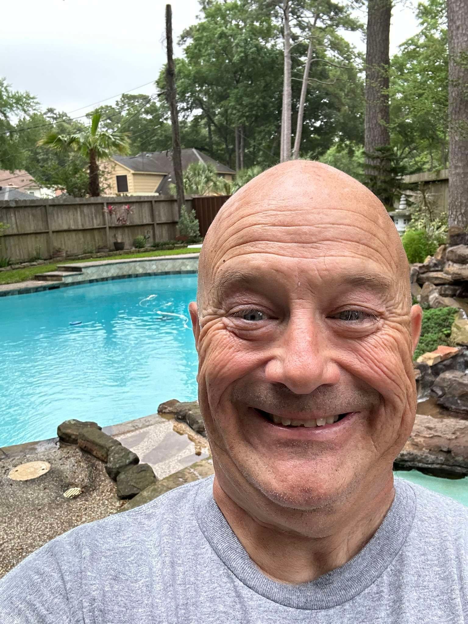 Bald man smiles in front of a blue swimming pool. Gray shirt, outdoors, trees in background.