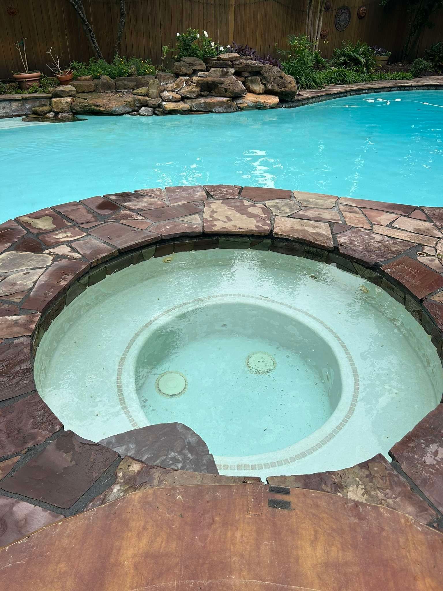 A circular spa built into a patio with a swimming pool behind it.