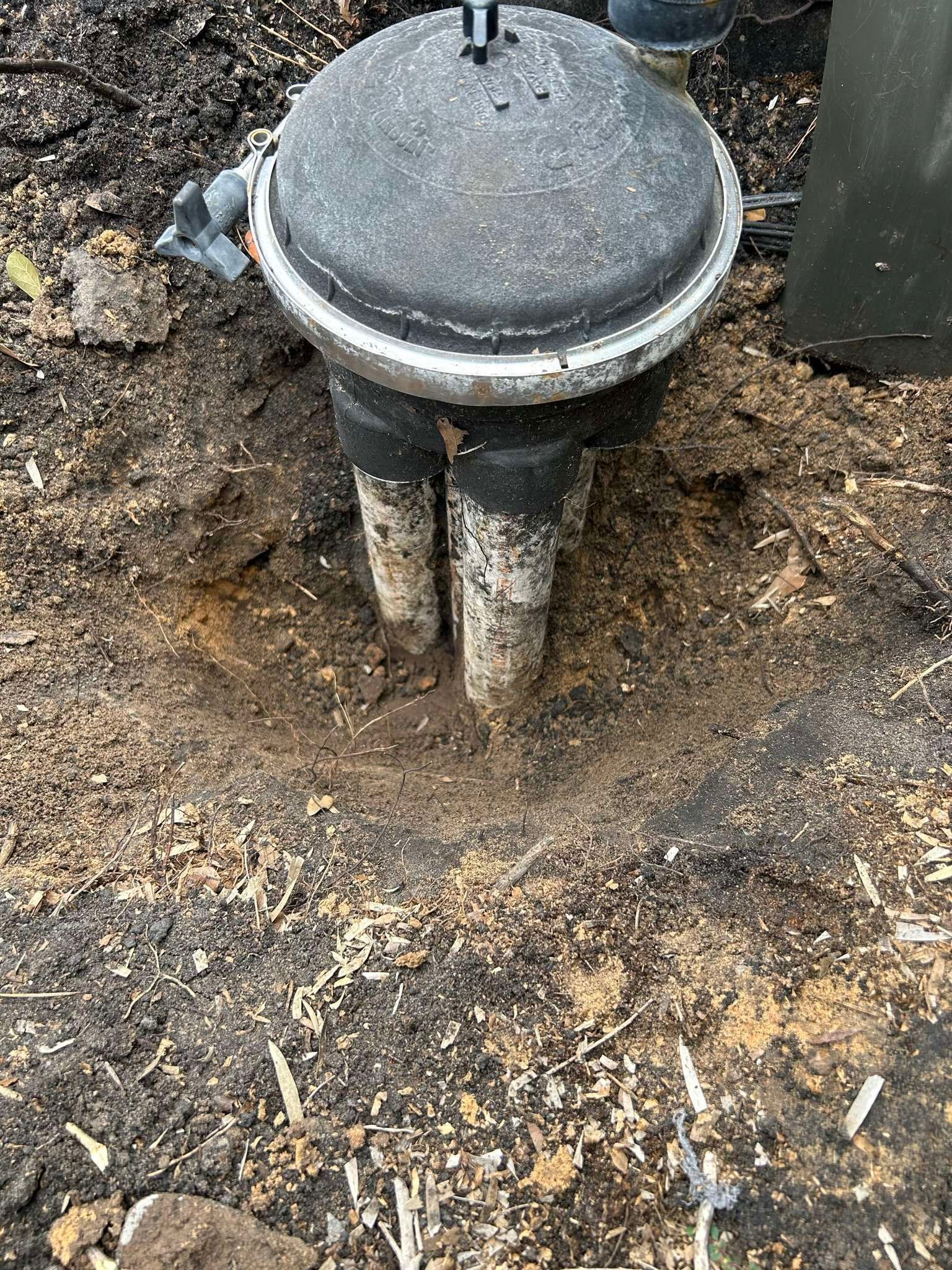 Black, cylindrical outdoor well with two pipes emerging from the ground, surrounded by dirt and mulch.