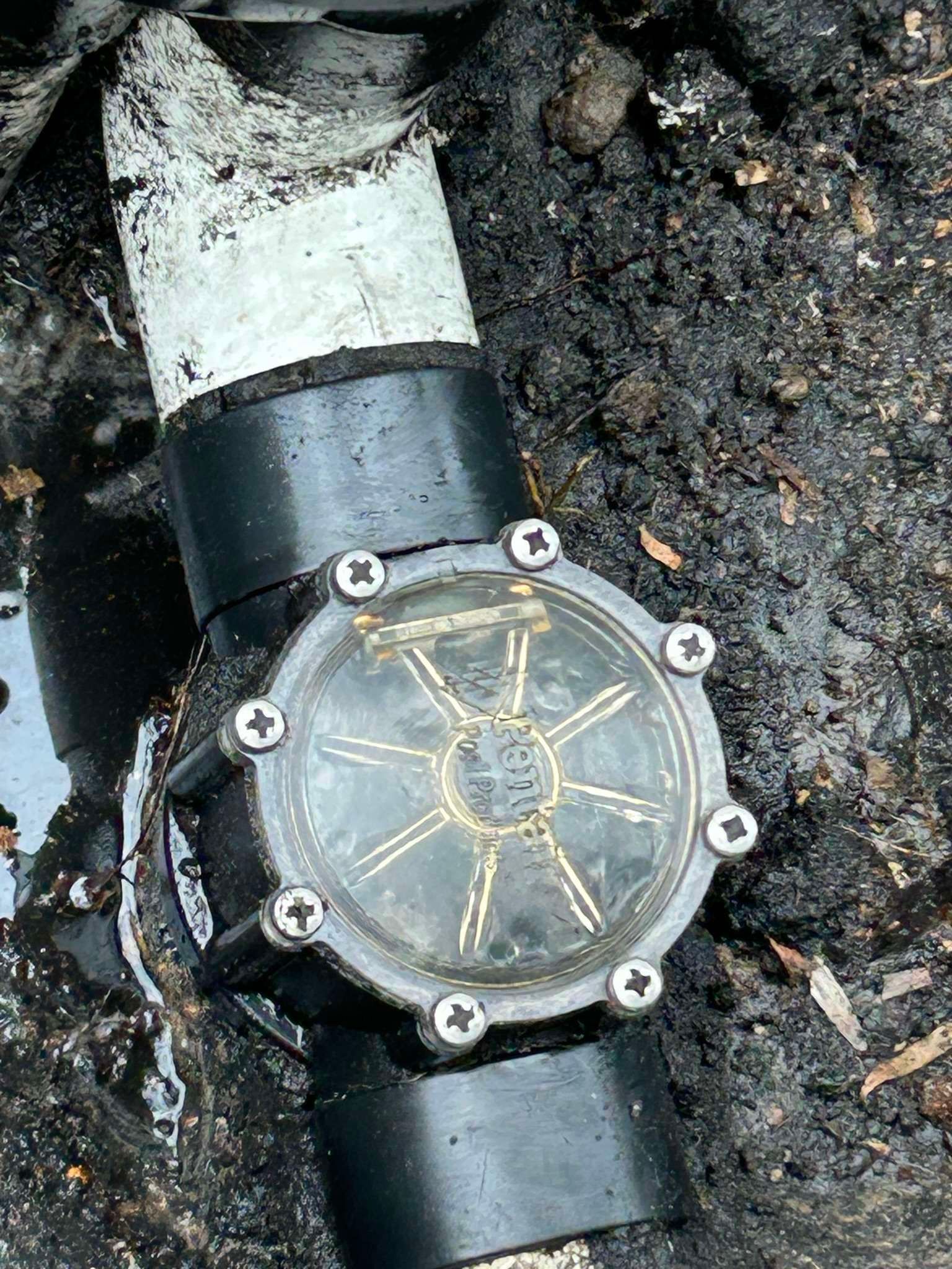 Black plastic pipe with a clear circular inspection port, surrounded by soil.