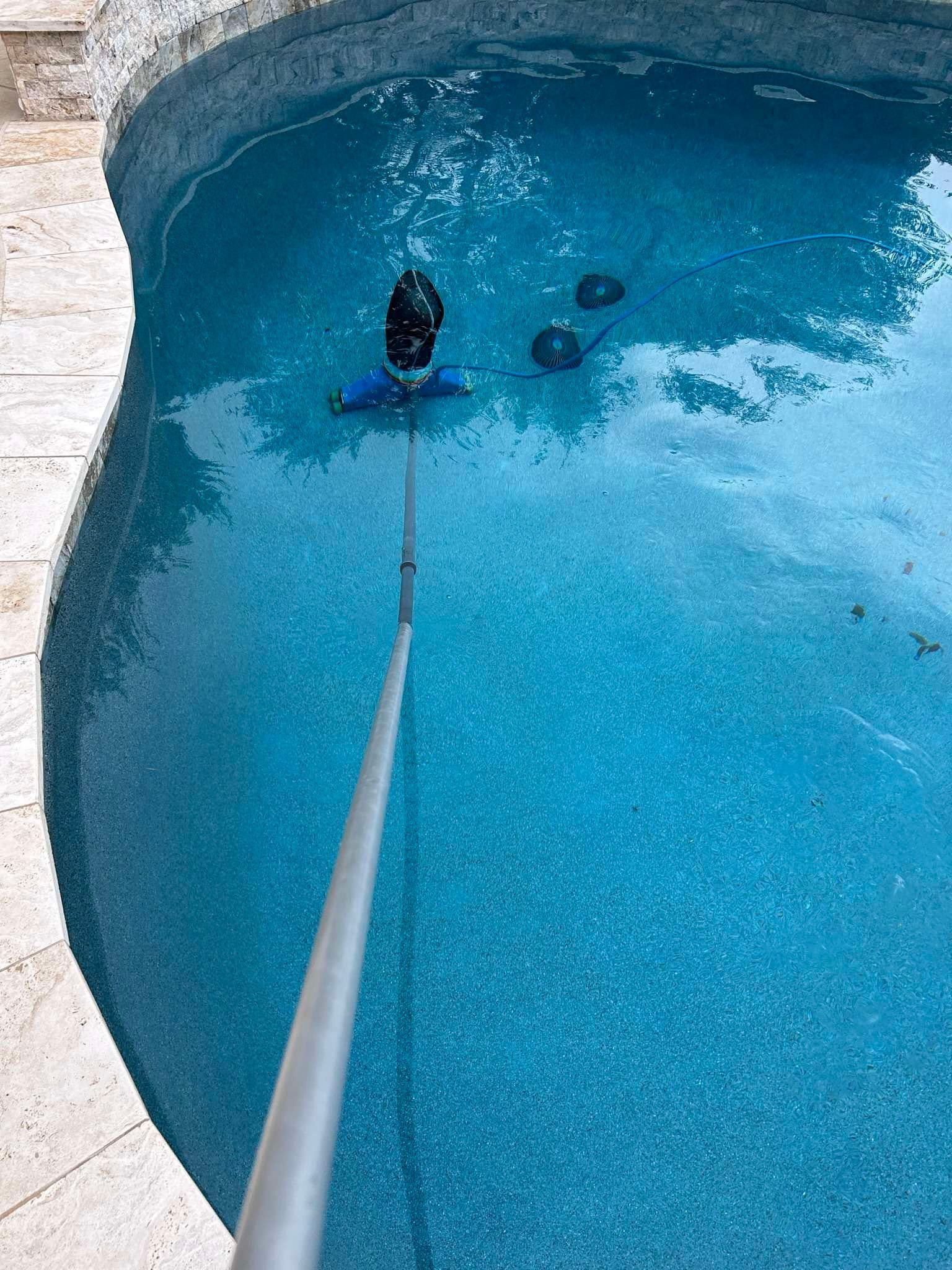Pool being cleaned with a blue vacuum head on a long pole; clear blue water.