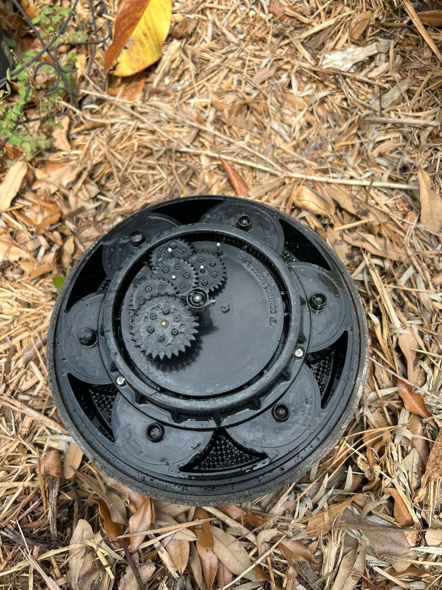 Black sprinkler head with exposed gears, sitting in mulch and dried leaves.