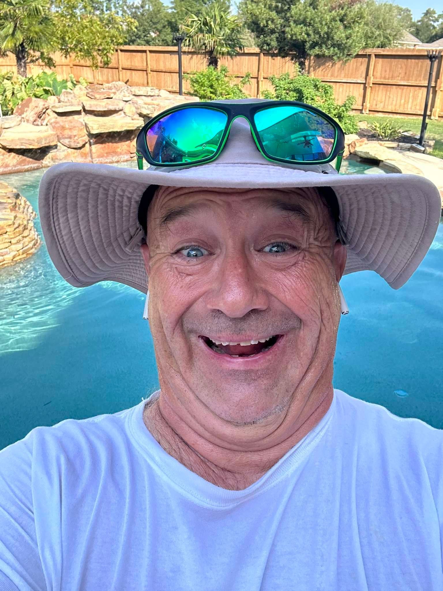 Man in hat with sunglasses smiles by a pool.