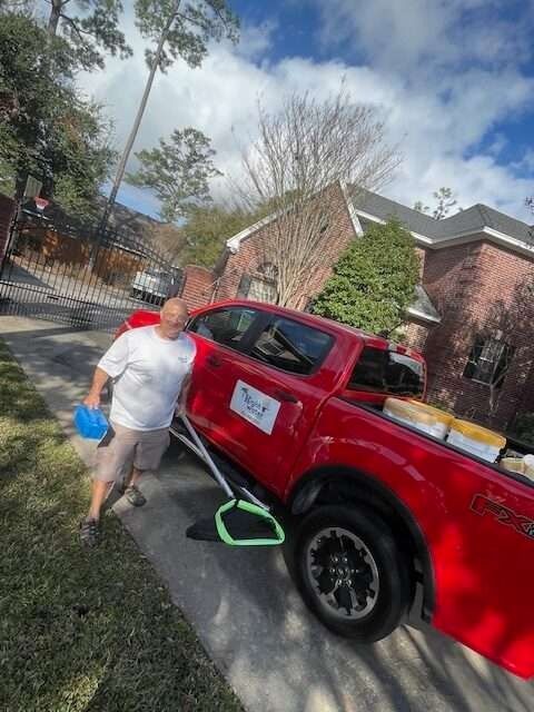 Man next to red pickup truck, parked in front of brick house. He is holding a blue hard hat and smiling.