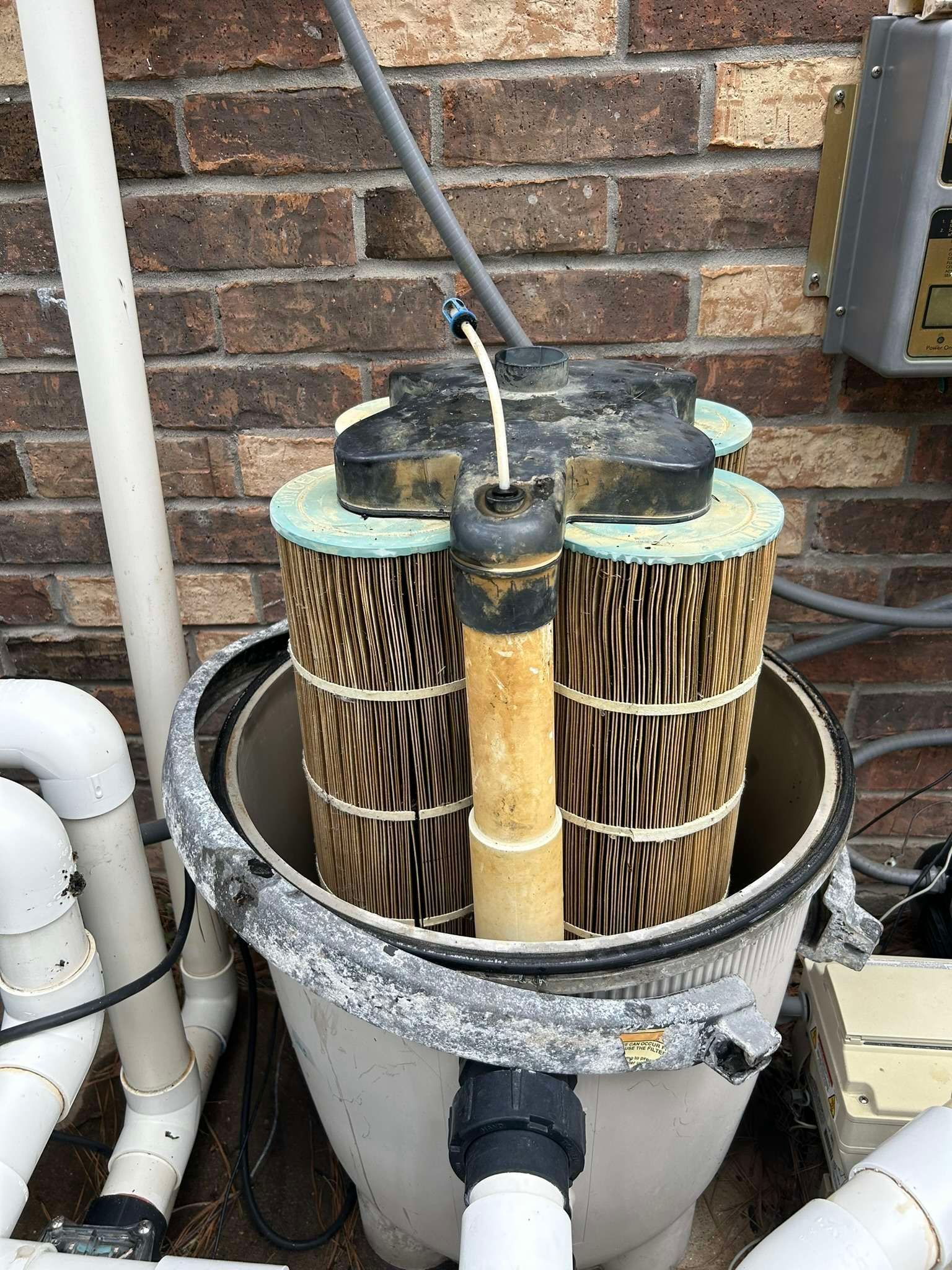 Pool filter with three cartridge filters inside a gray tank, near a brick wall.