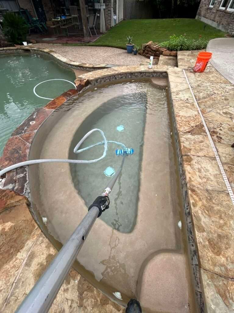 A person cleaning a spa with a vacuum attached to a pole. The spa is next to a pool.