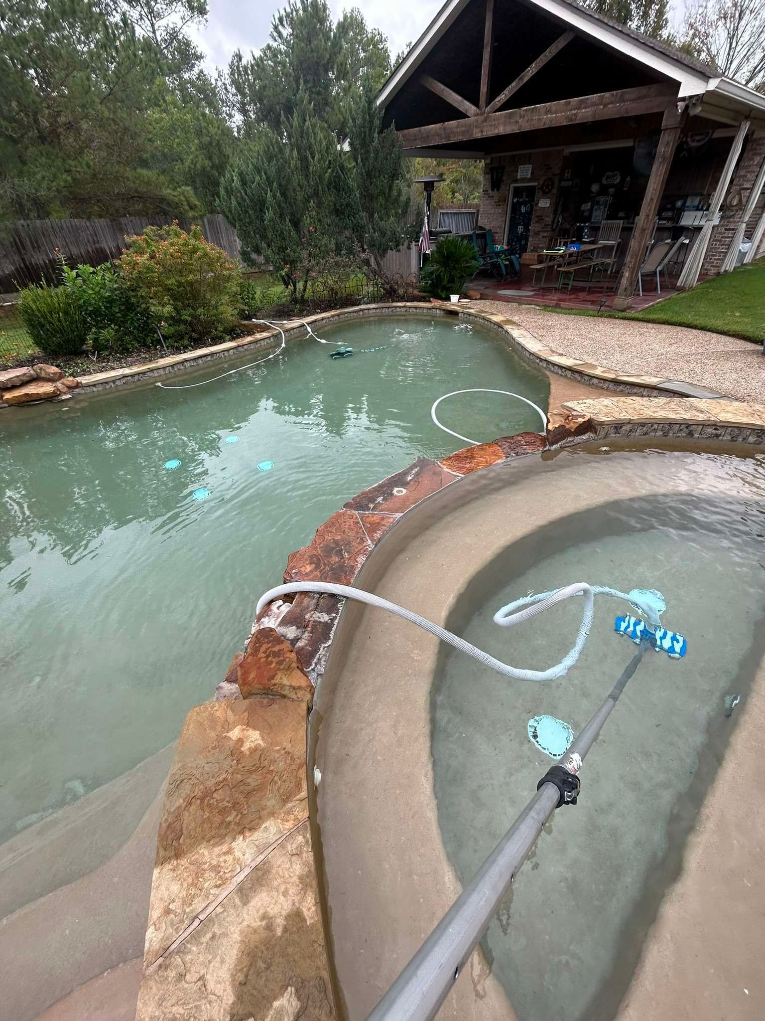 Pool being cleaned with a blue scrubber in a light green pool and a hot tub. A covered patio sits in the background.