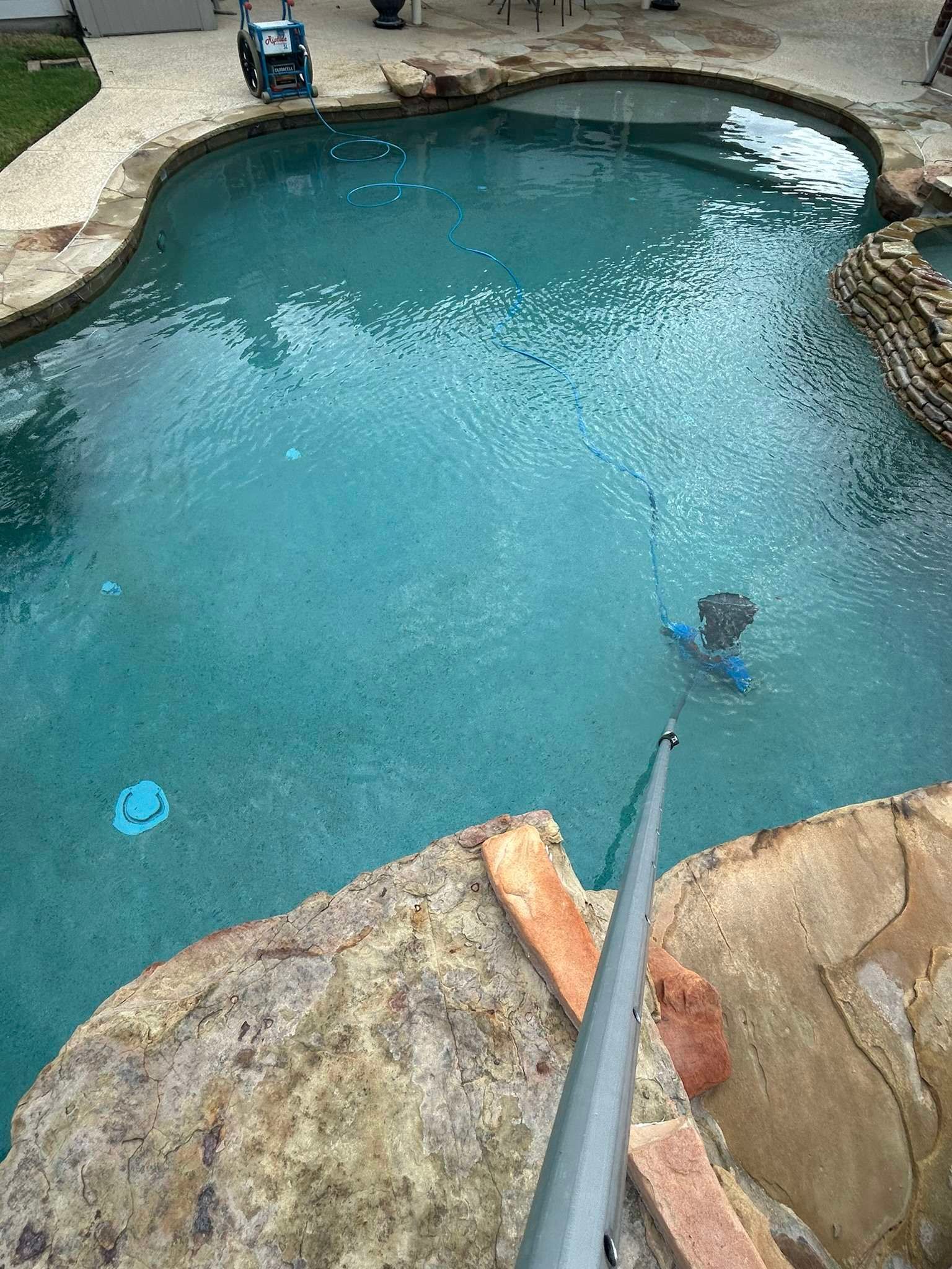 Person cleaning a turquoise pool with a long-handled brush, stone coping around the edge.