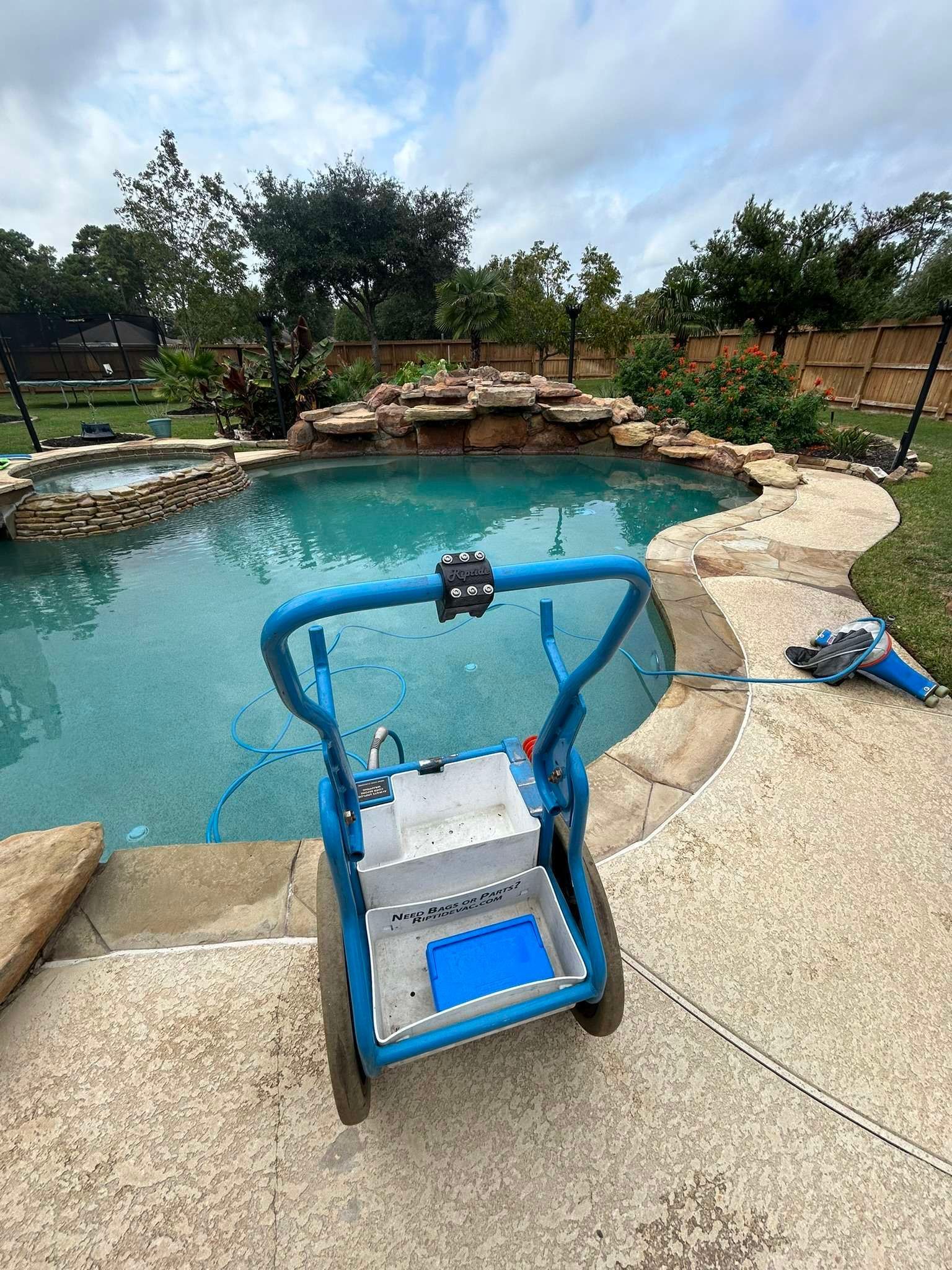 Blue pool cleaner near a pool with a waterfall in a backyard.