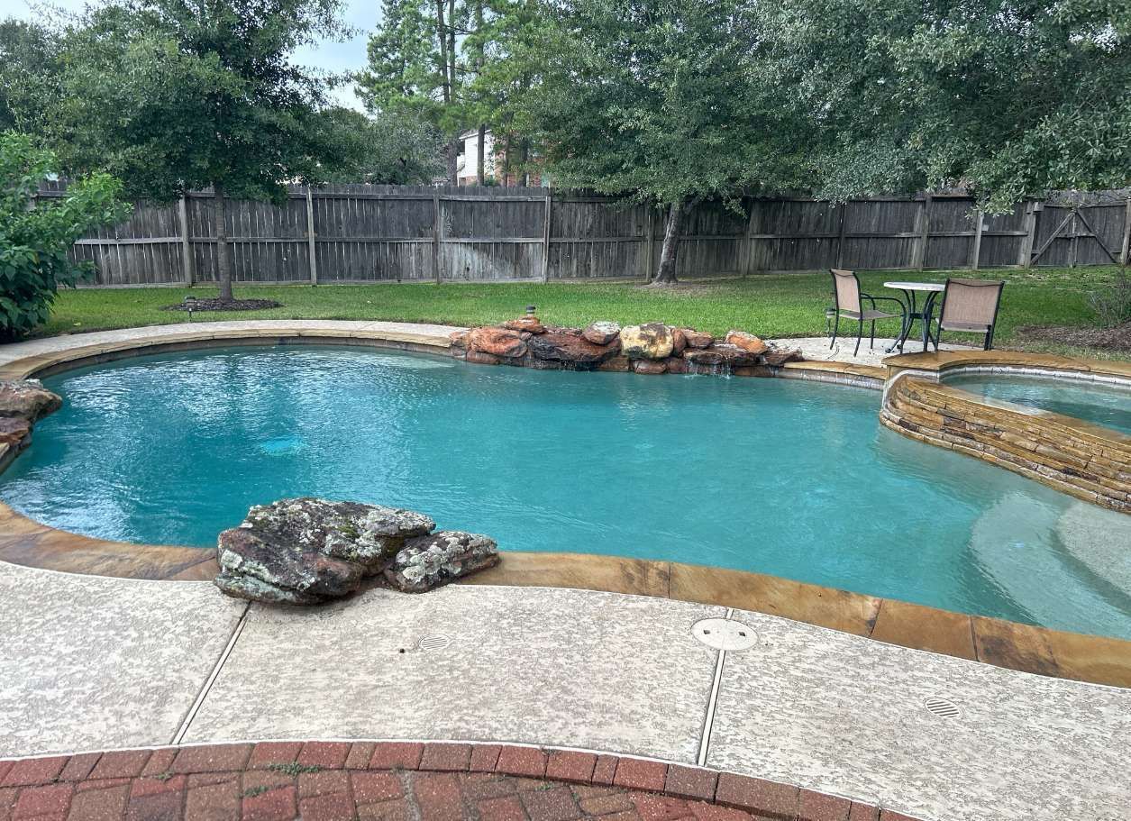 A backyard pool with rock features and seating area. Green water, concrete patio, and wooden fence.