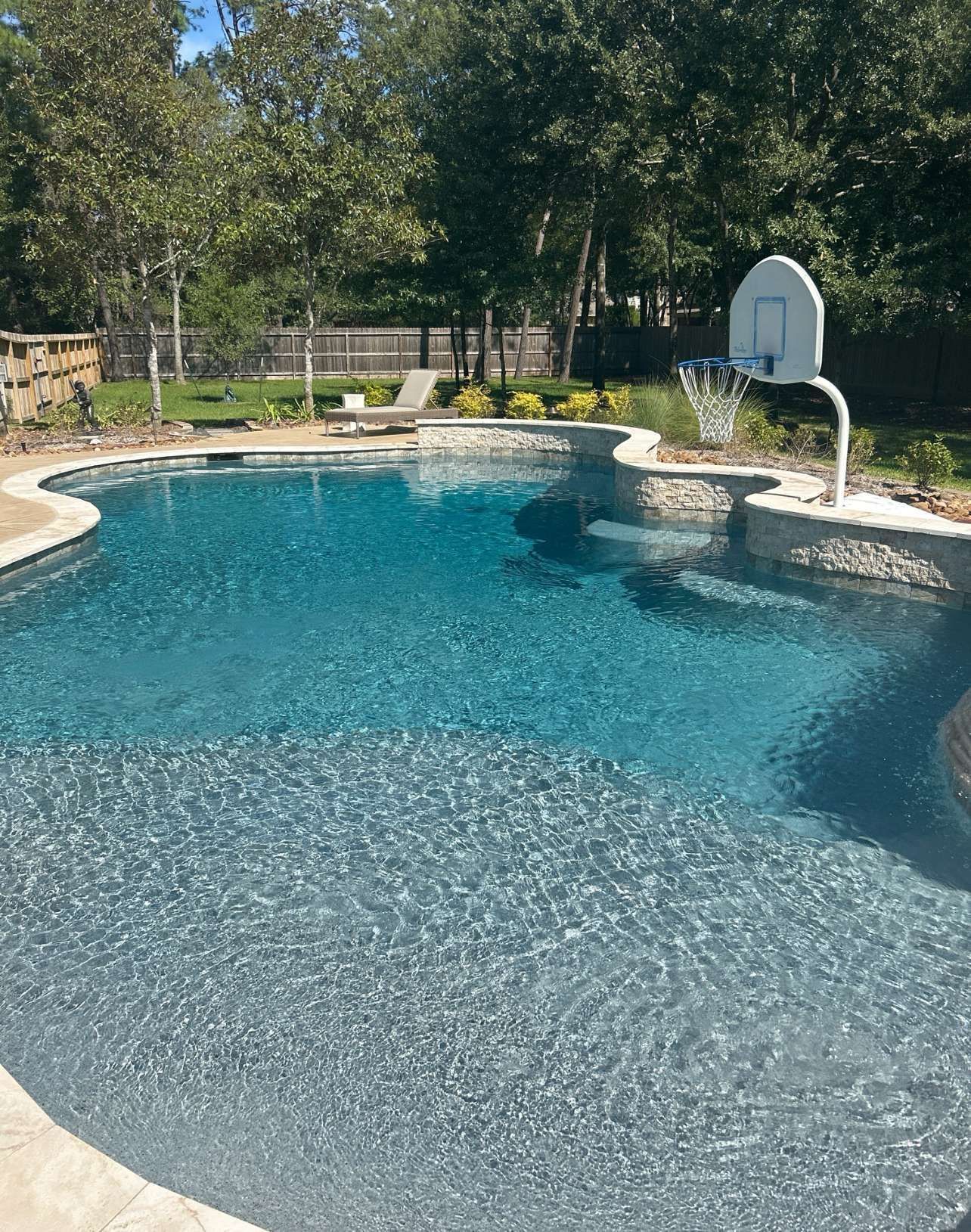 Blue swimming pool with basketball hoop, stone surround, and trees in the background.