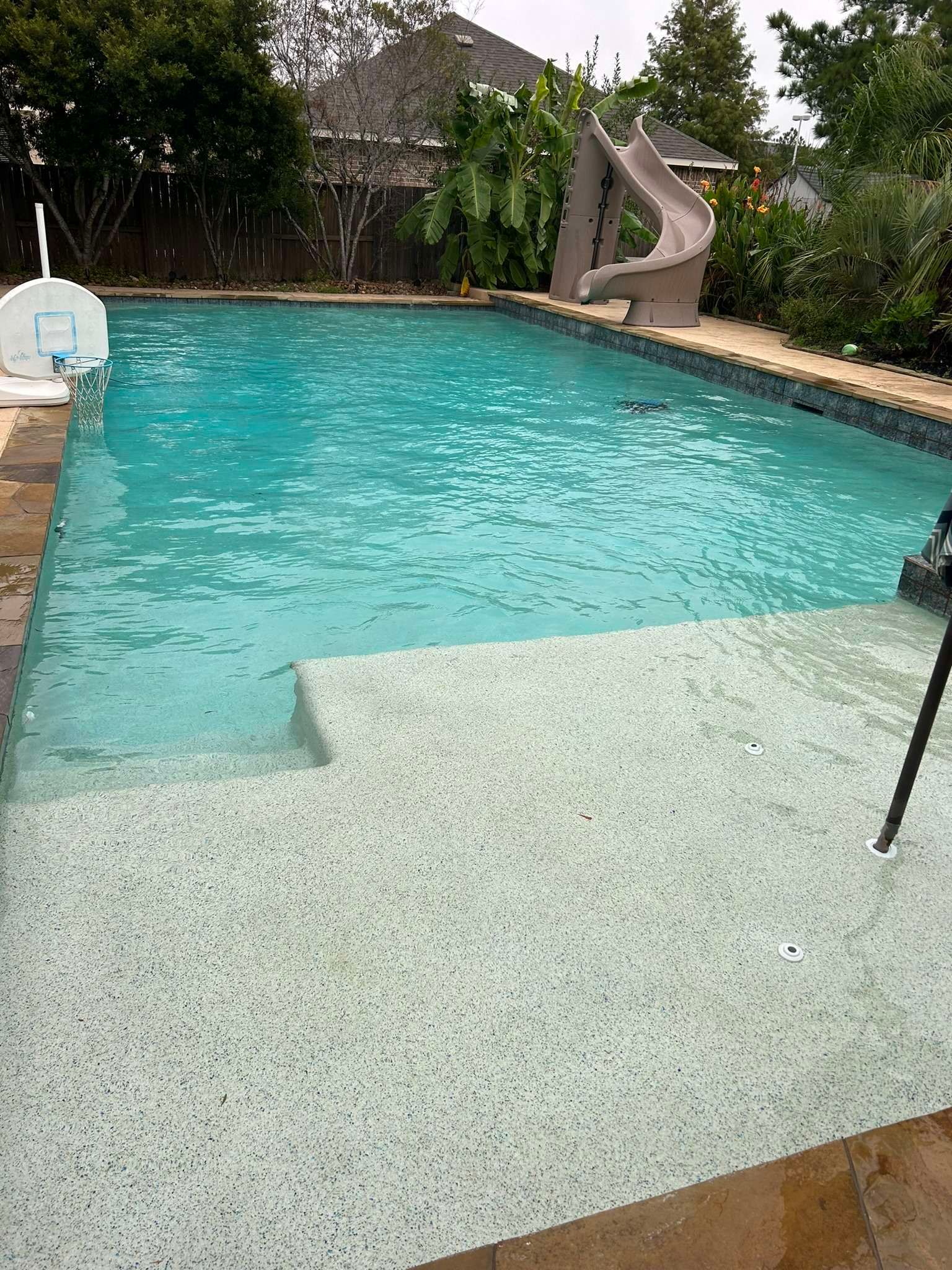 A rectangular swimming pool with built-in steps and a slide in a backyard setting.