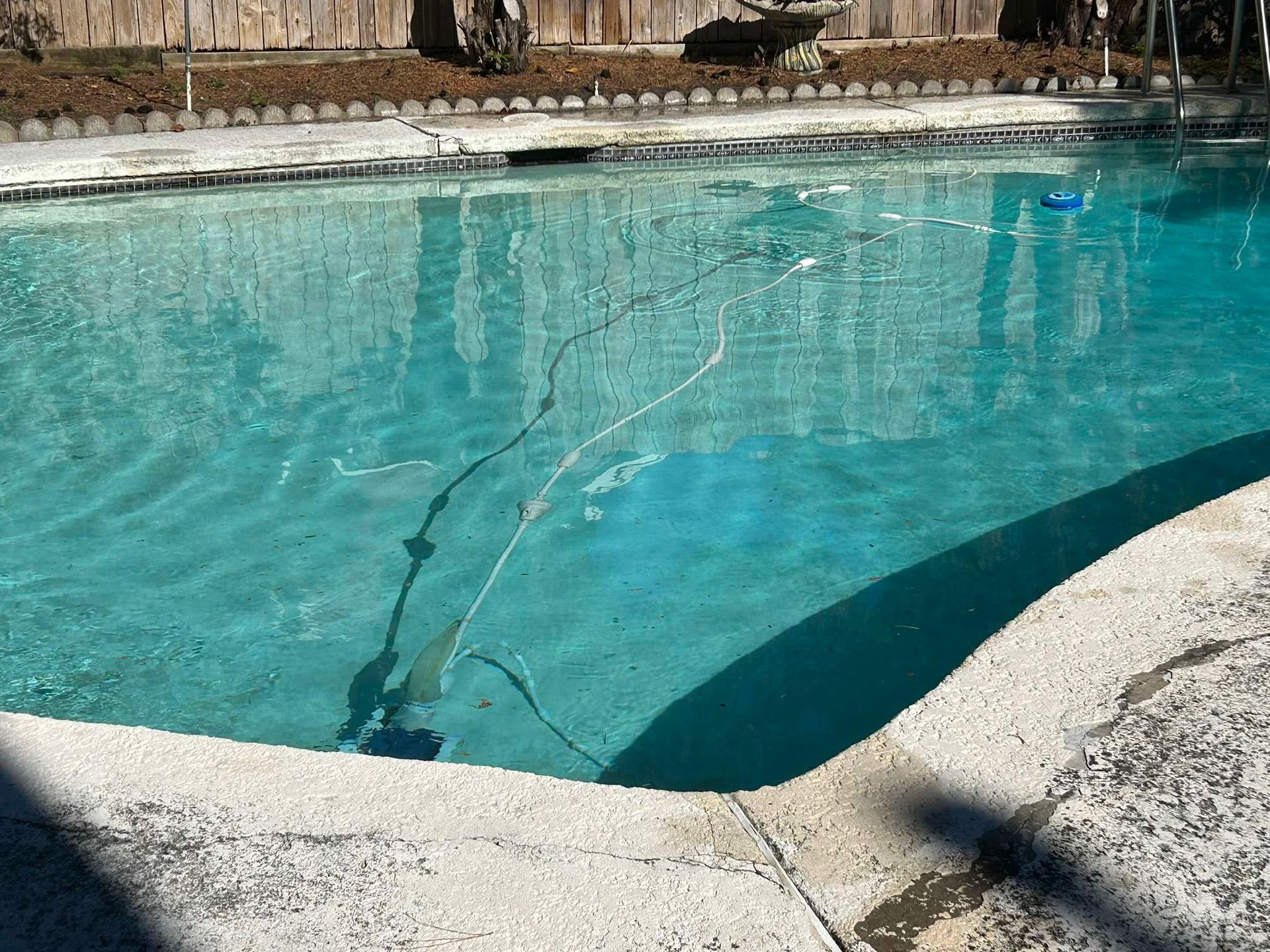 A swimming pool with clear water, a pool cleaner submerged, surrounded by a concrete deck and a wooden fence.