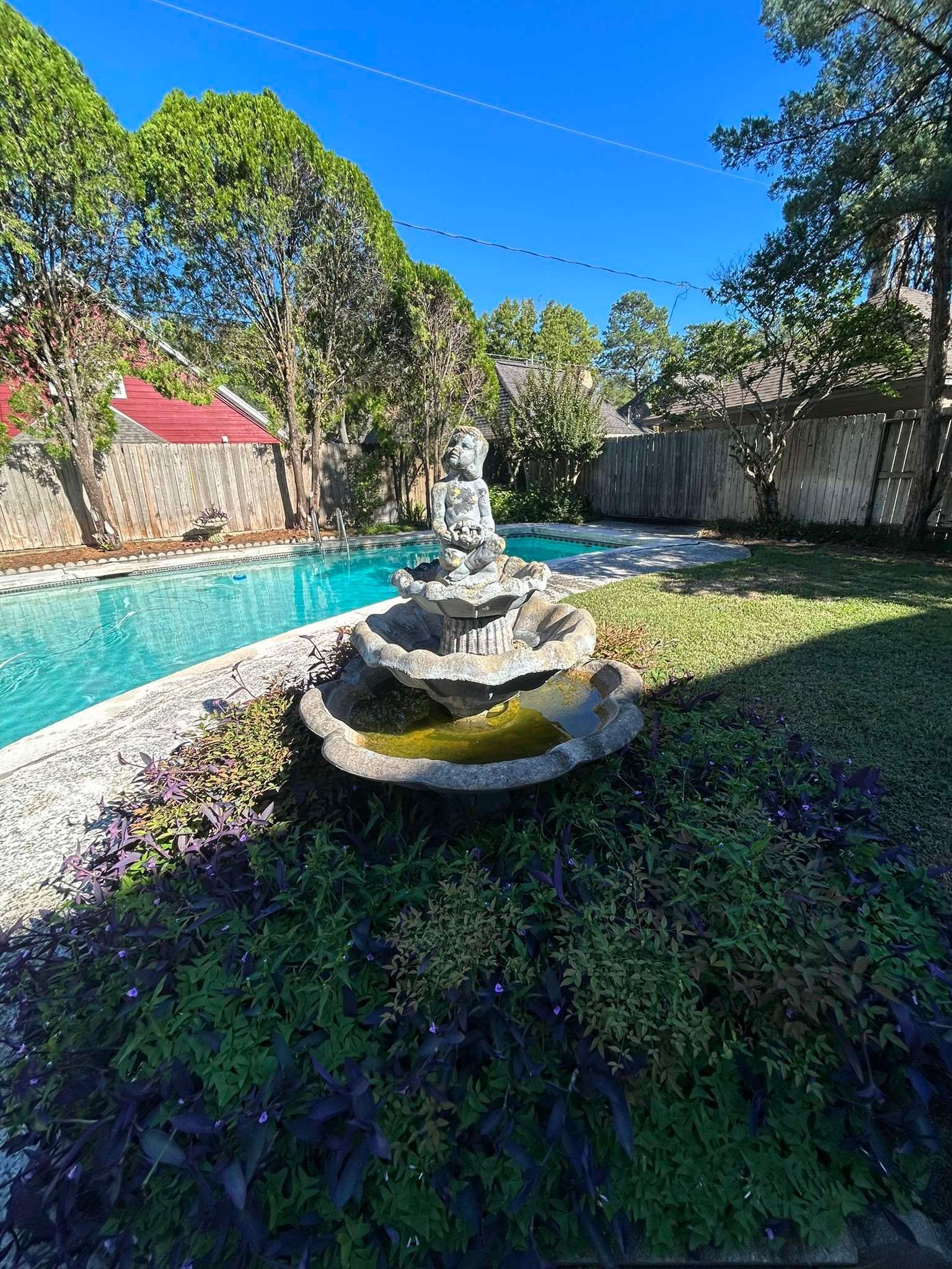 A serene backyard with a pool, fountain statue, and lush greenery under a bright blue sky.