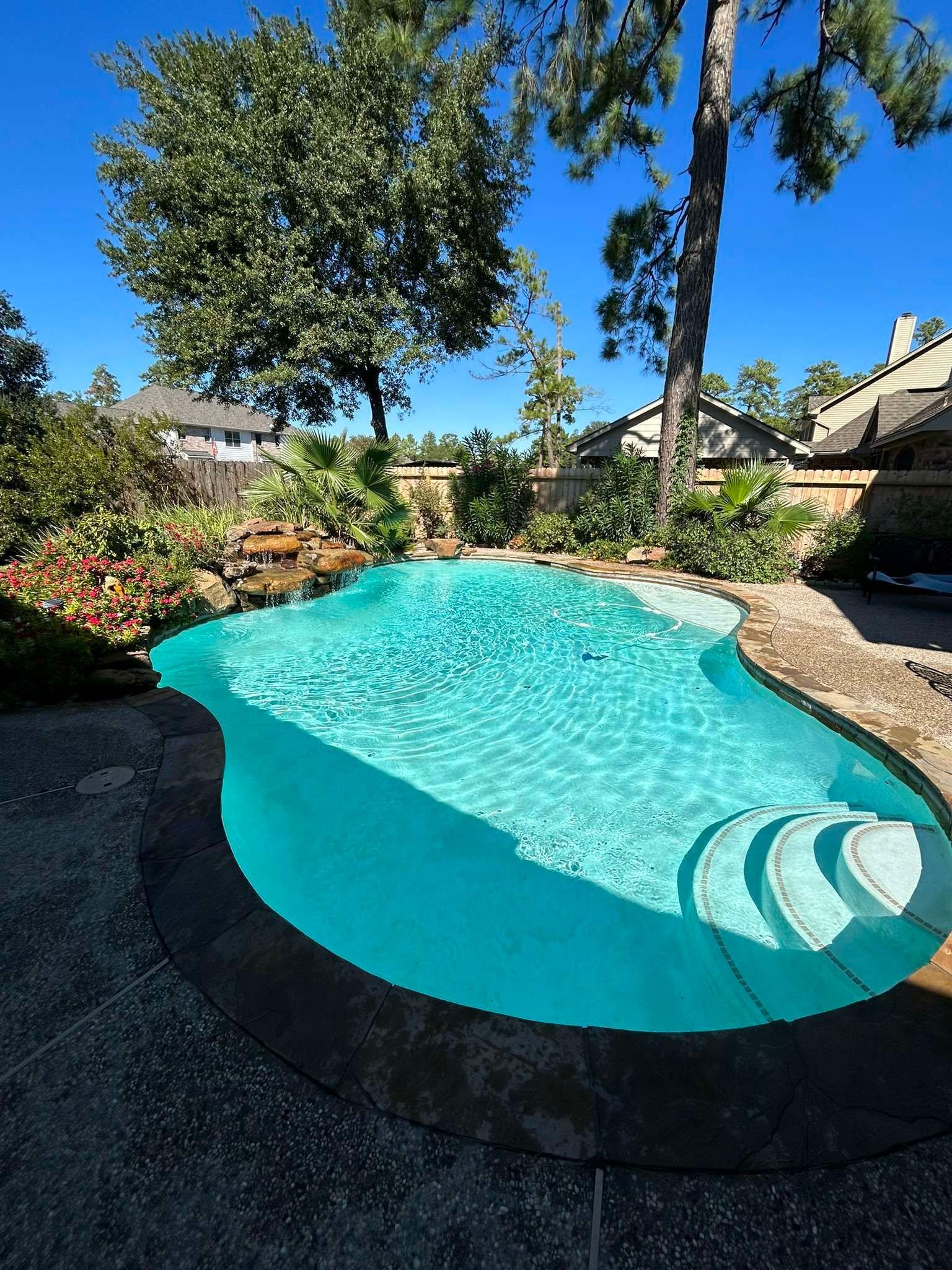 Swimming pool with clear turquoise water, surrounded by stone patio and landscaping, under a blue sky.