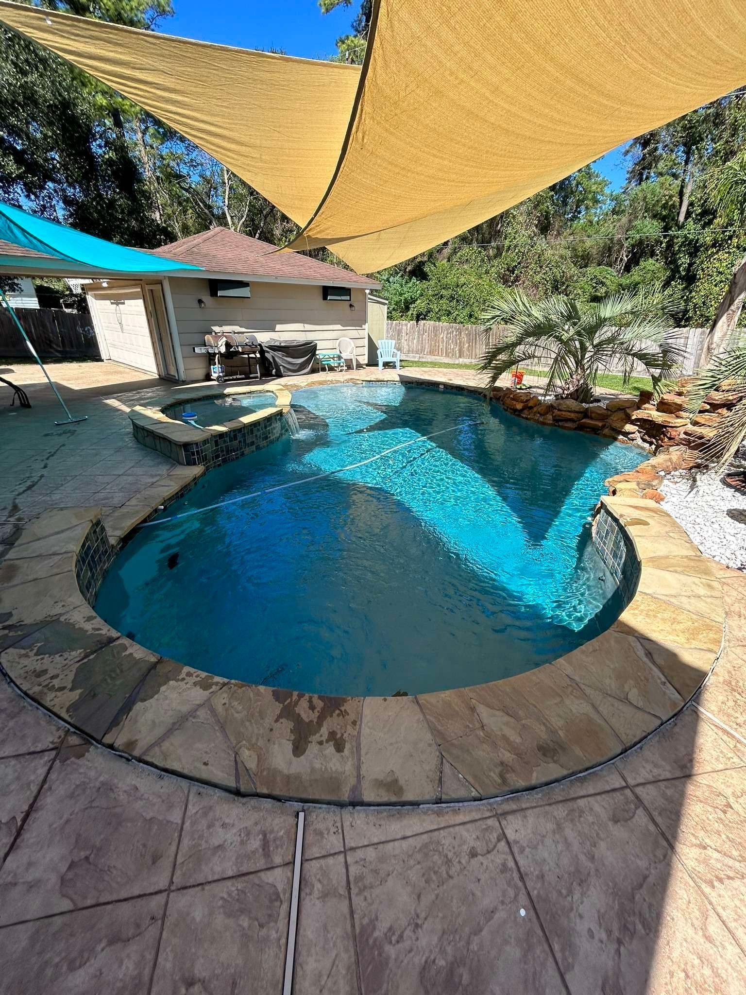 A pool with a tan shade cover. The pool is surrounded by stone and includes a jacuzzi, waterfall, and small building.