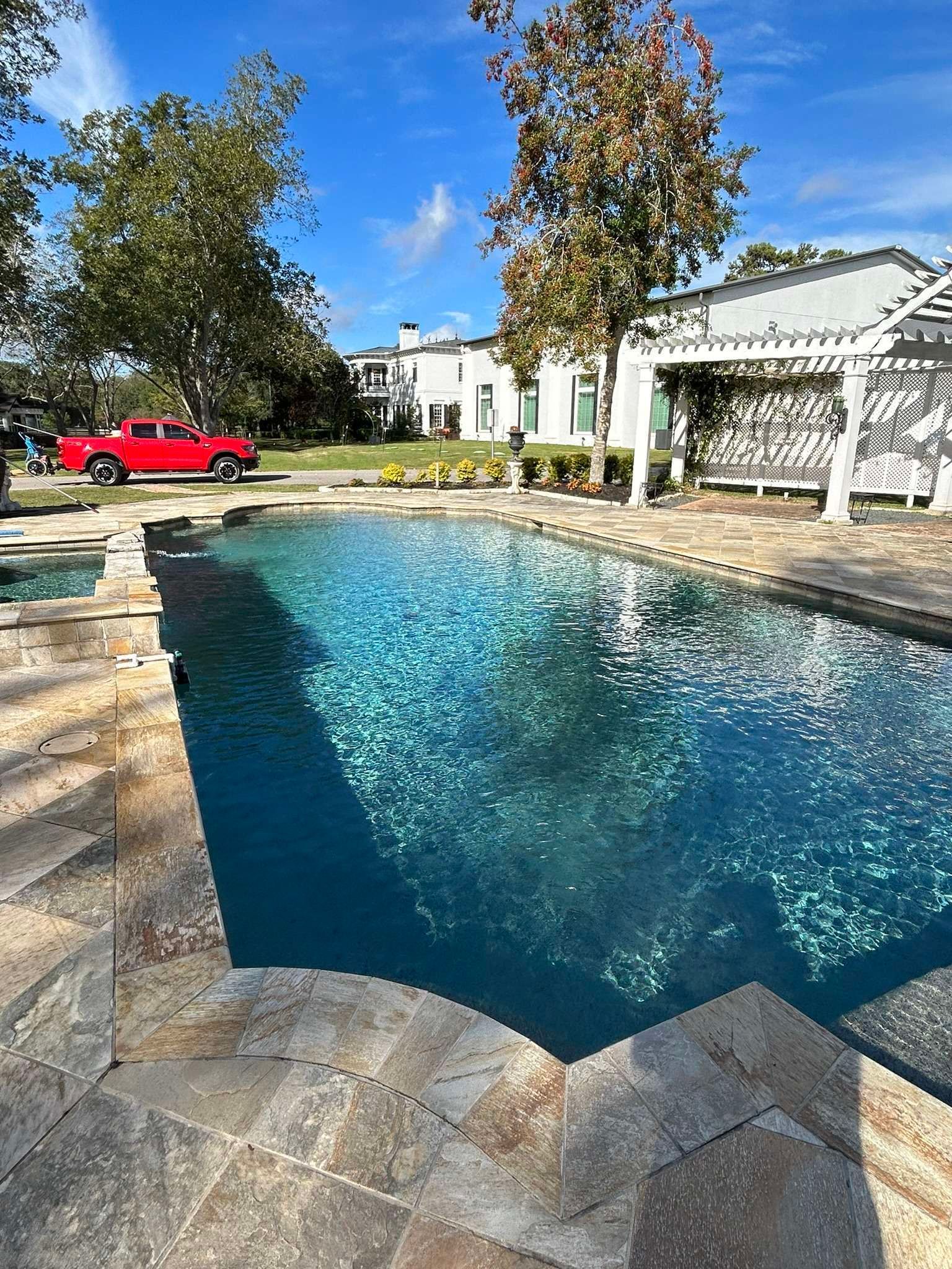 Swimming pool with blue water and stone surround, near a white pergola and a red truck.