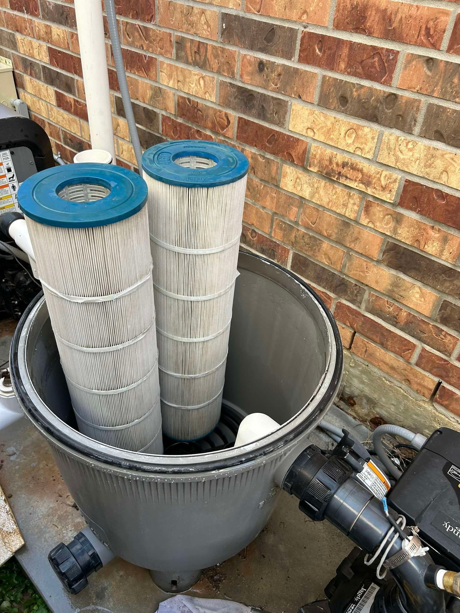 Pool filter housing with two tall, cylindrical filter cartridges. Brick wall in the background.