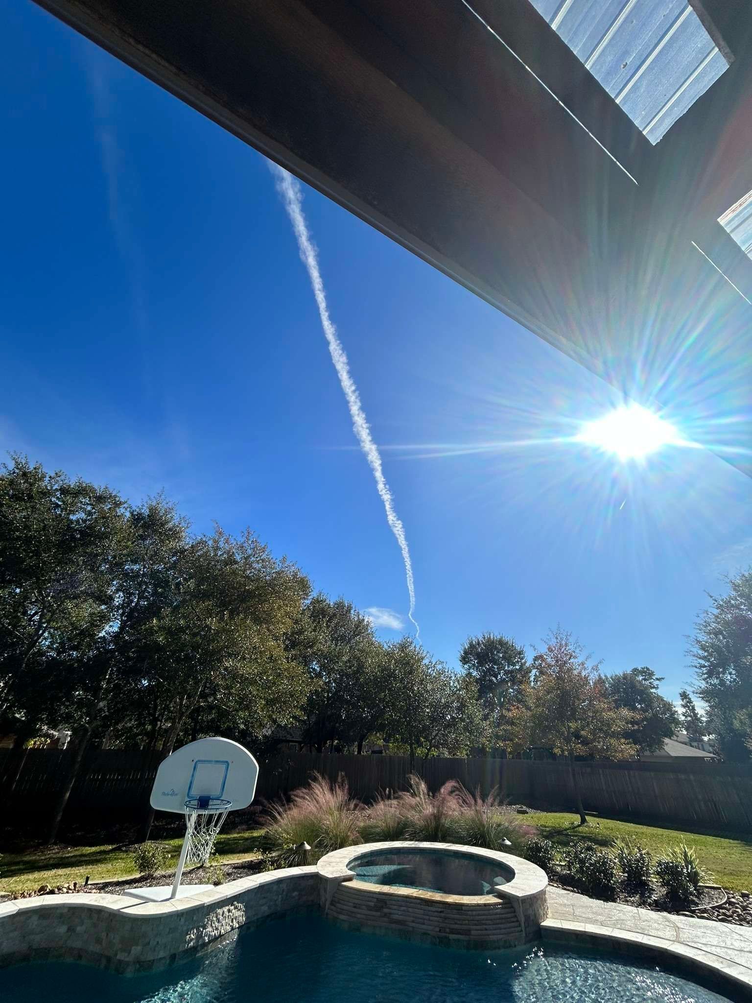 Poolside view with bright sun, blue sky, and a contrail. Trees and a basketball hoop are visible.