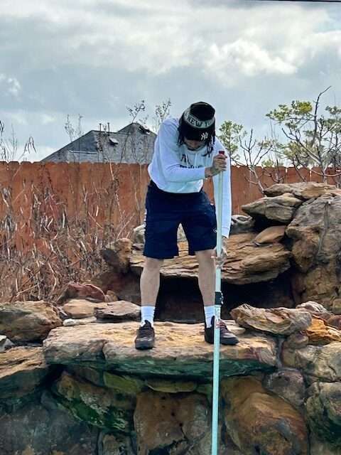 Person leans over a rock pond, holding a long pole. Cloudy sky, brown fence in background.