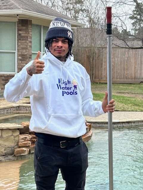 Man in white hoodie with thumbs up, holding a pool cleaning pole. Next to a pool.