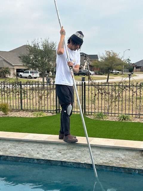 Person cleaning a pool with a long pole, standing near a pool's edge, sunny outdoor setting.