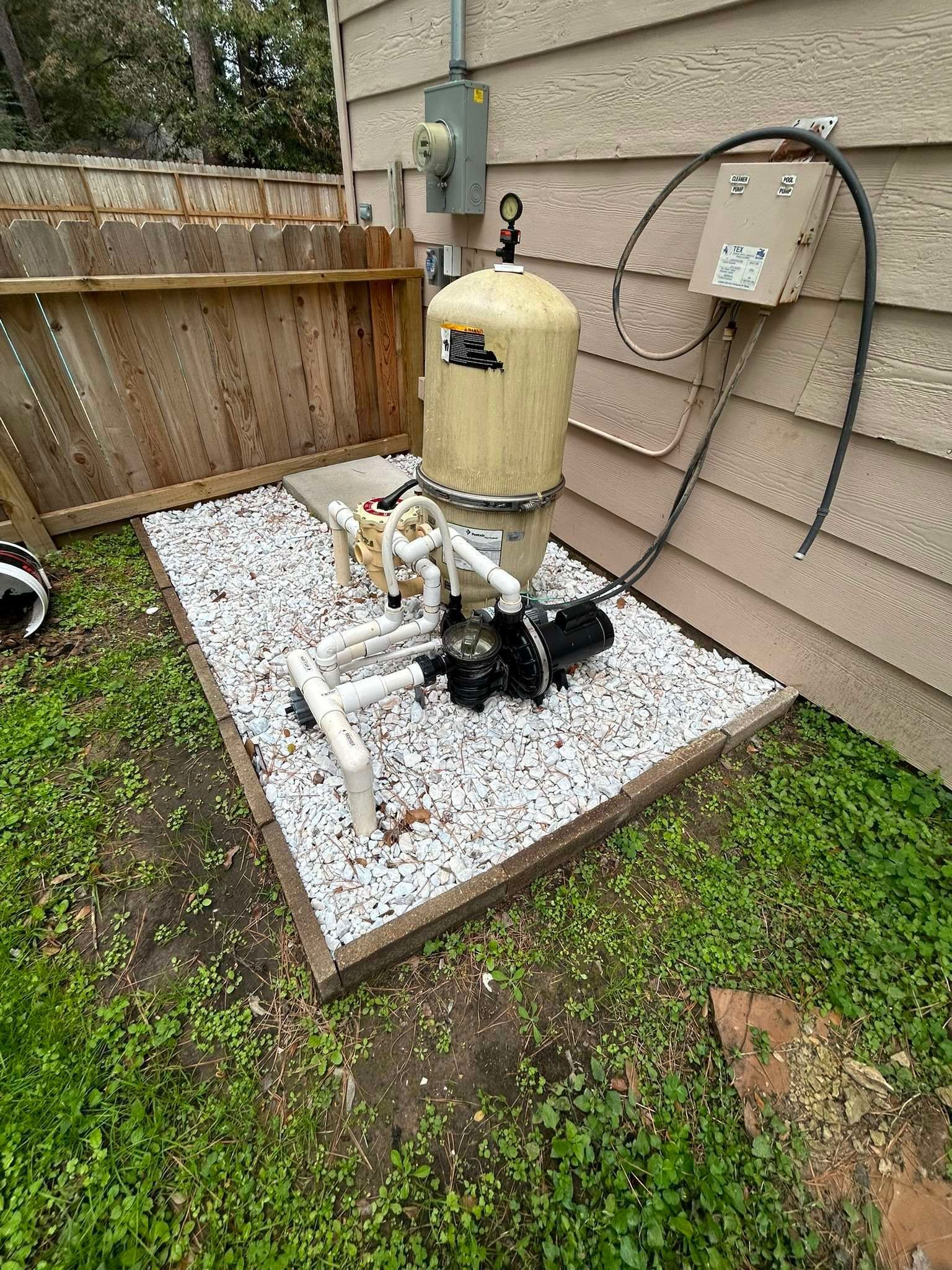Pool filter system with a beige tank and black pump on a bed of white stones next to a wood fence.