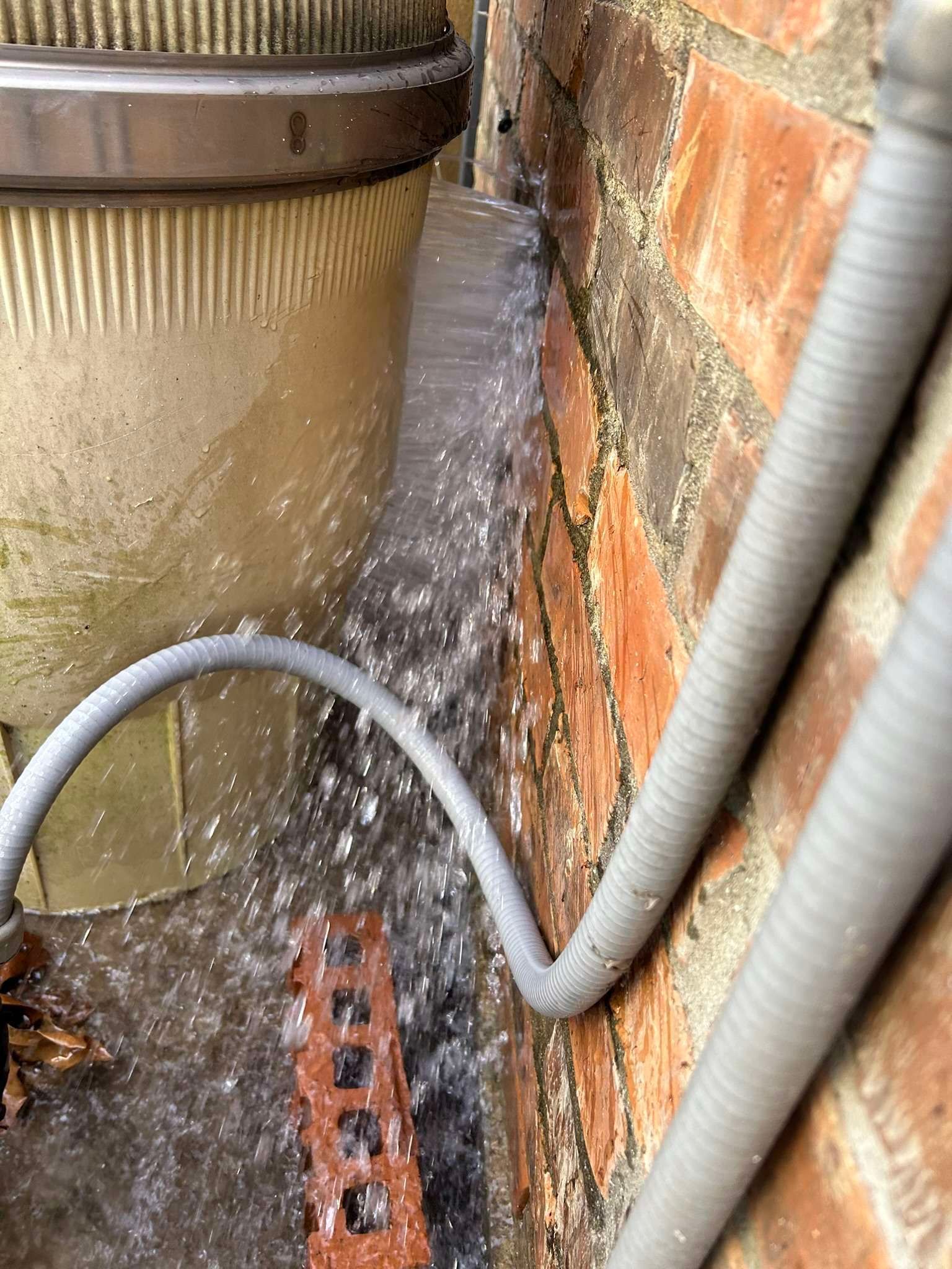 Water gushing from a light-colored cylinder near a brick wall, splashing onto a brick and ground.