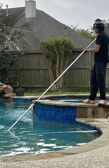 Person using a long pole to clean a pool; outdoors, overcast day.