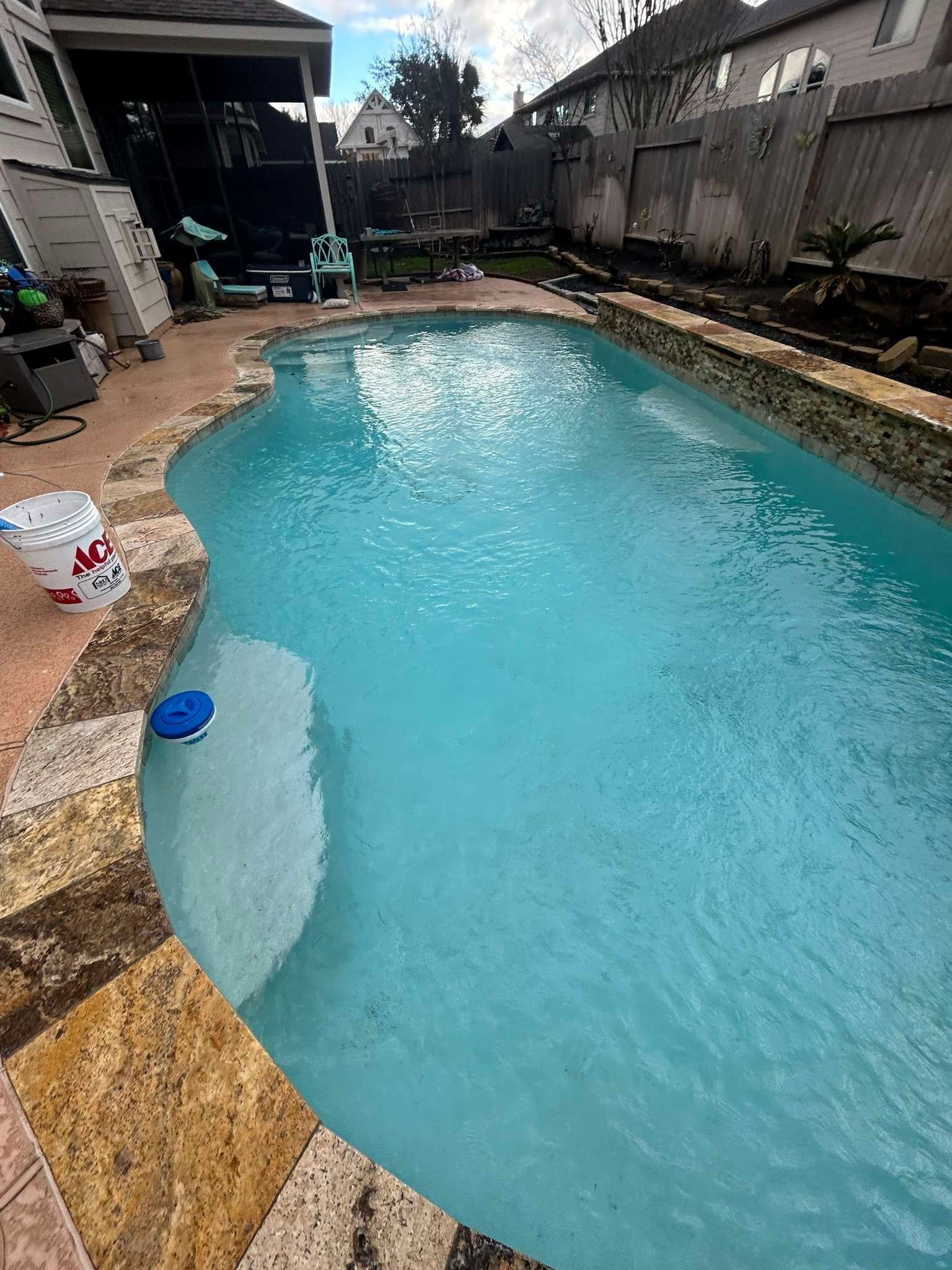 Swimming pool with turquoise water, surrounded by stone and a backyard patio.
