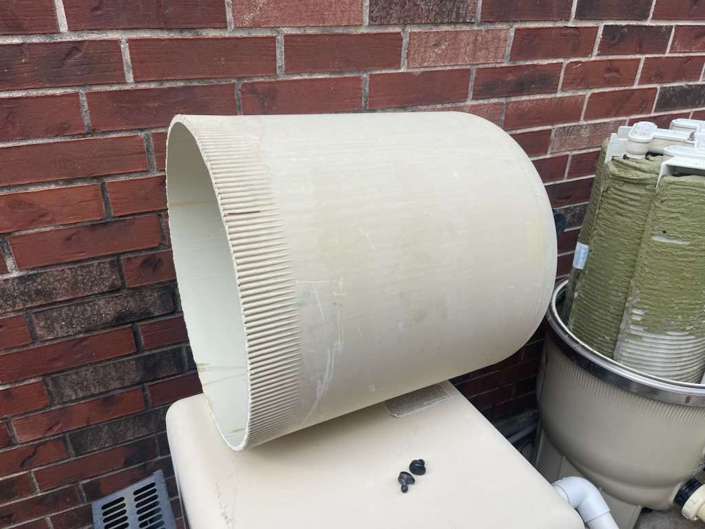 Large, off-white pool filter cartridge component, laying on top of a filter housing, near a brick wall.
