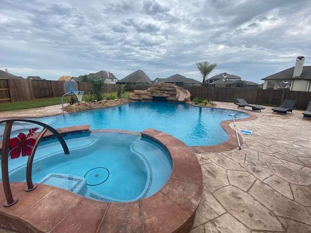 Backyard pool with spa, waterfall, slide, and lounging area under a cloudy sky.