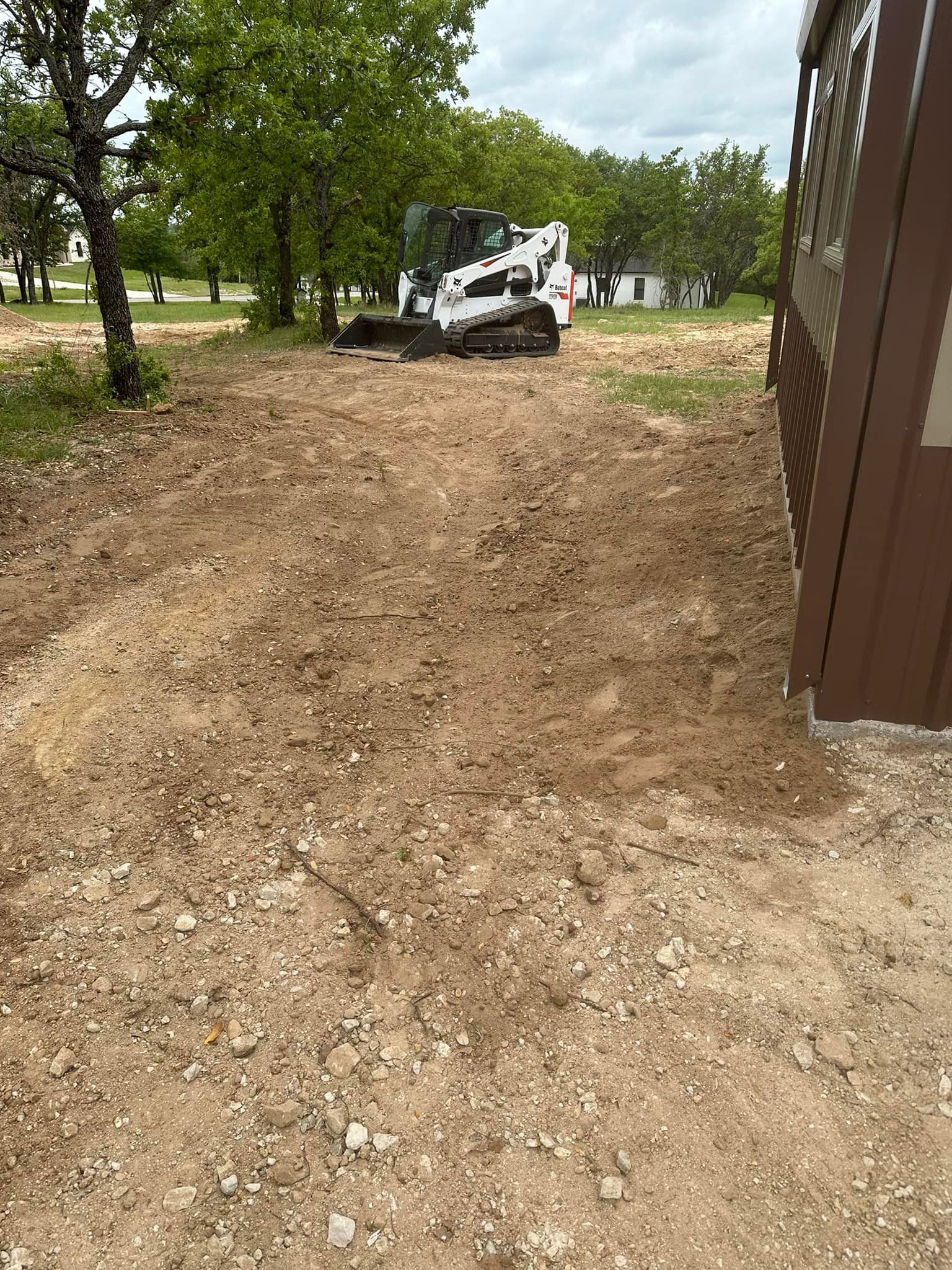 A bobcat is driving through a dirt field next to a shed.