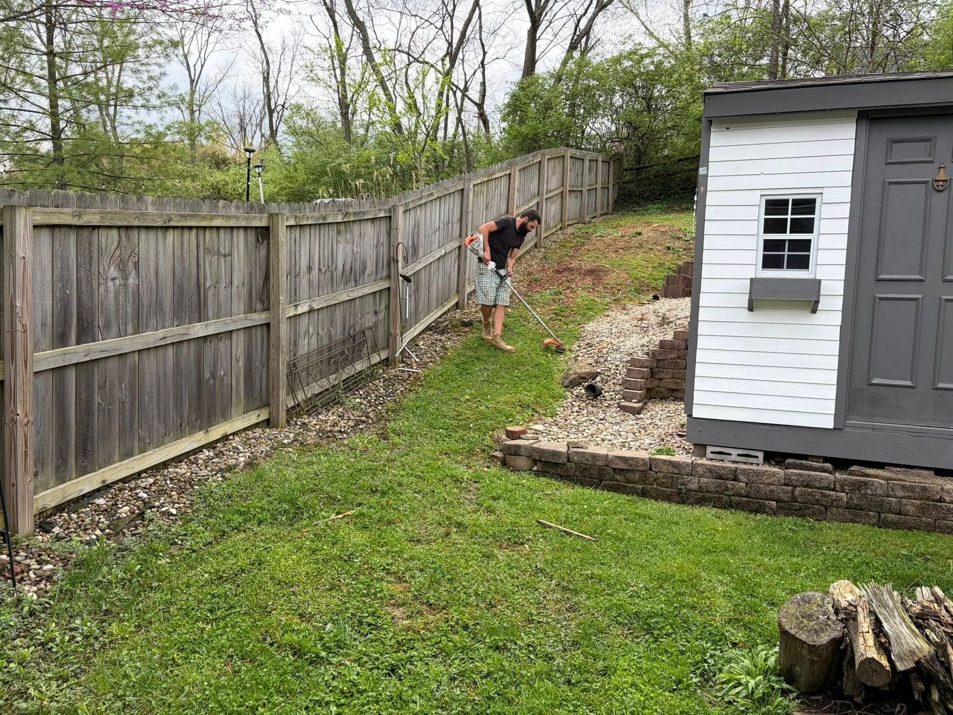 Person trimming a sloped backyard along a wooden fence beside a small shed.
