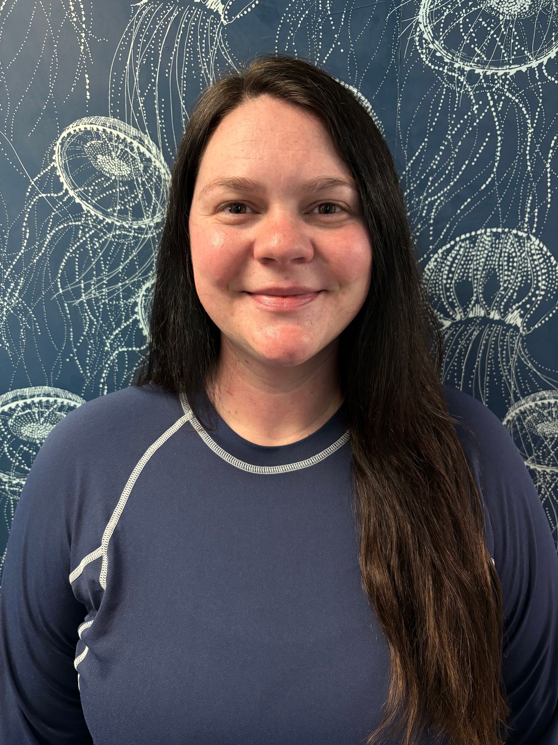 A woman in a purple shirt is smiling in front of a wall with jellyfish on it