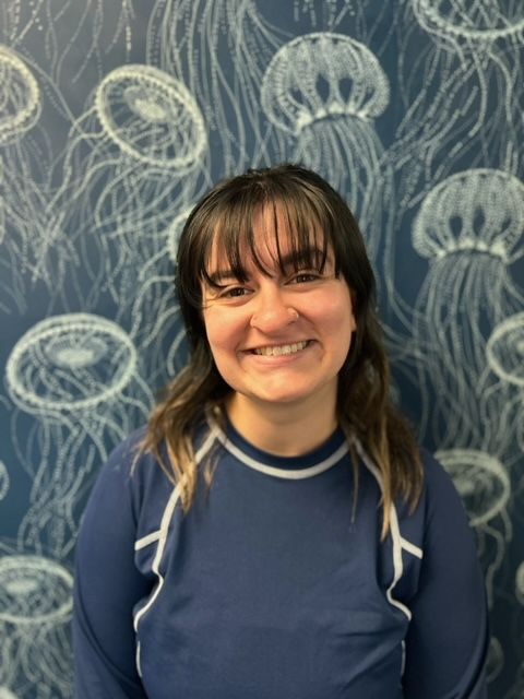 A woman in a blue shirt is smiling in front of a wall with jellyfish on it.