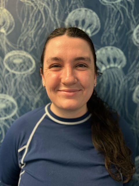 A woman in a blue shirt is smiling in front of a wall with jellyfish on it.