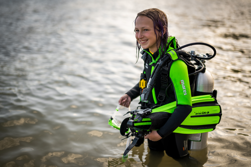 A young girl in a green scuba suit is kneeling in the water.