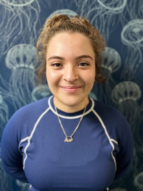 A woman wearing a blue shirt and a necklace is smiling in front of a jellyfish wallpaper.