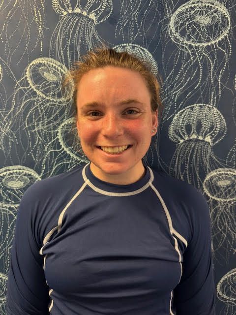 A woman in a blue shirt is smiling in front of a wall with jellyfish on it.