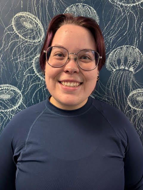A woman in a blue shirt is smiling in front of a wall with jellyfish on it