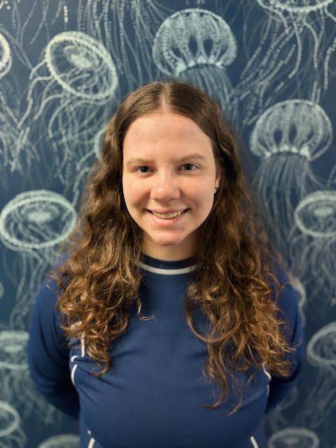 A woman in a blue shirt is smiling in front of a wall with jellyfish on it.