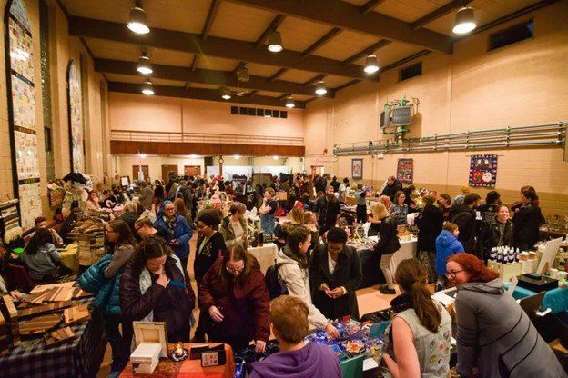 Photo of a warm toned hall with many vendor tables and patrons walking around with a busy and bustling energy